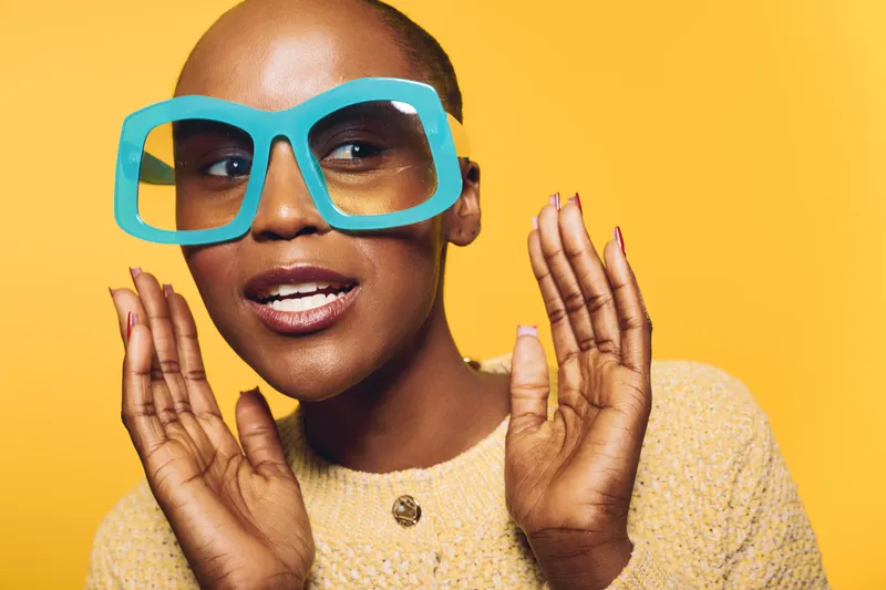 Close-up of a confident black woman with dark skin wearing oversized turquoise glasses and a beige sweater, posing against a bright yellow background. Concept of fashion and expression.