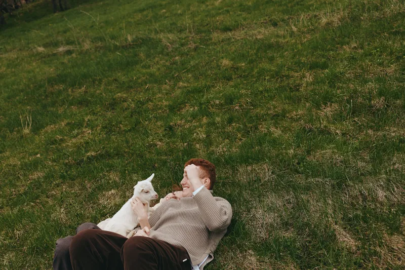 Cheerful man relaxing outdoors with a curious goat kid beside him, sunlit grass, casual clothing, warm smile, peaceful moment, nature setting, candid lifestyle scene