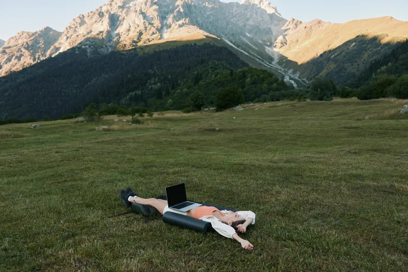 A person lies on a grassy hillside with a laptop, enjoying a peaceful outdoor work session in a mountainous landscape, sunlight bathes rolling terrain and distant peaks.