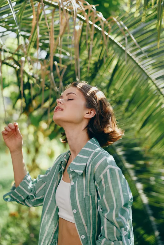 A woman stands with eyes closed, basking in warm sunlight amid tropical foliage, wearing a striped green shirt and white crop top.