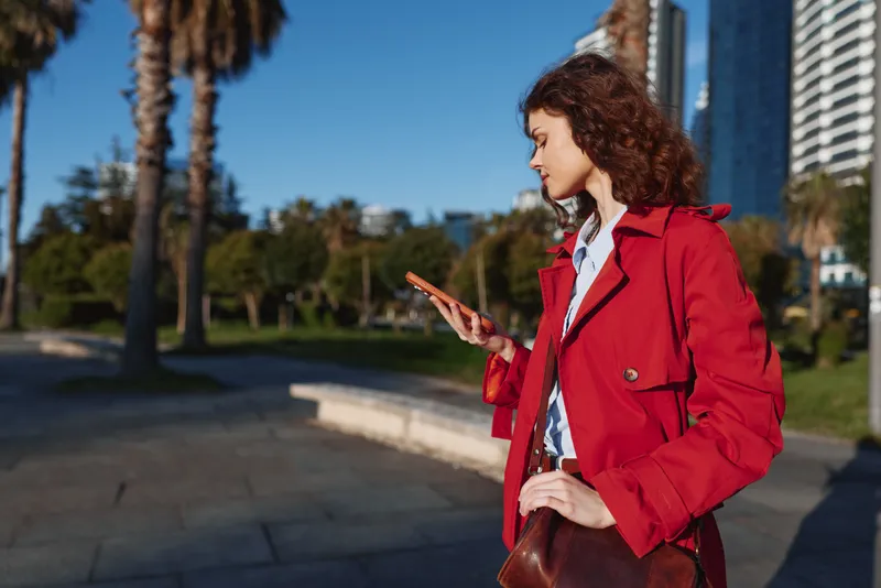 Woman in a red coat outdoors using a smartphone in a sunny park with palm trees and urban skyline, showcasing casual fashion, everyday technology, candid street style, natural light.
