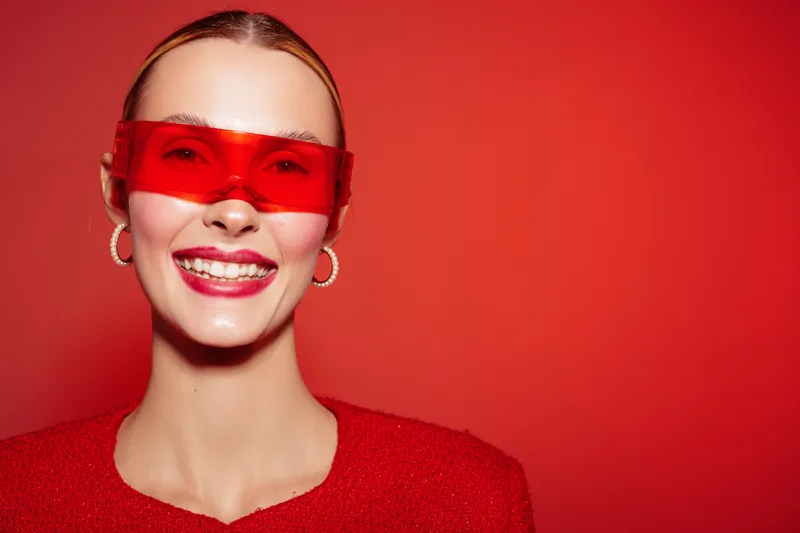 Smiling woman with red sunglasses and red outfit poses against red background. Bright makeup and hoop earrings create a bold, fashionable look with vibrant energy.