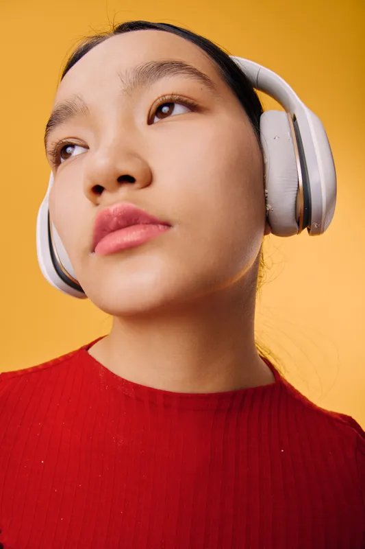 Young woman wearing headphones and red sweater listens to music with thoughtful expression on bright yellow background. Closeup portrait of female with clear skin and natural makeup.