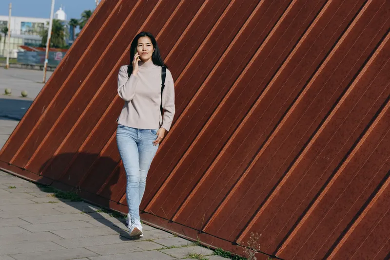 Woman in casual outfit stands beside a diagonal metal wall in an urban outdoor setting, showcasing street fashion and a confident pose with daylight and soft shadows.
