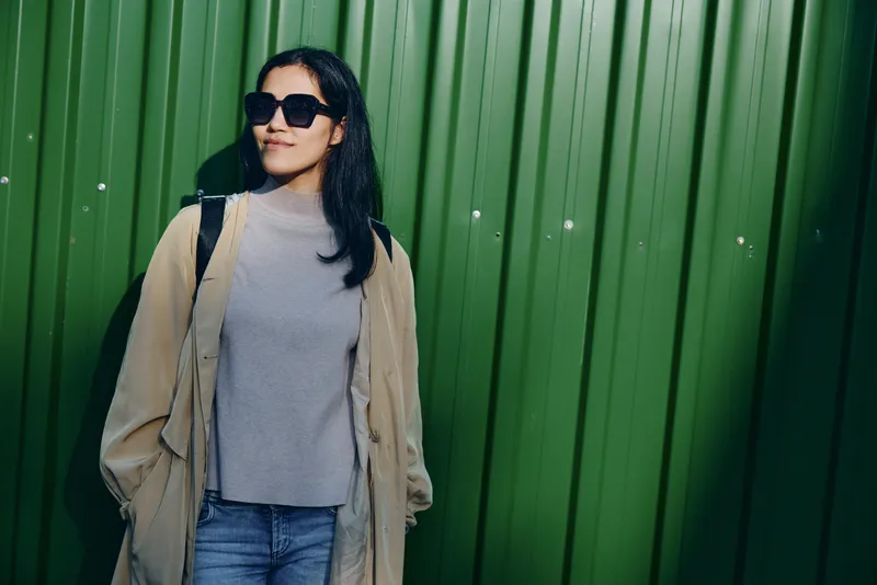 Woman wearing sunglasses and a beige coat leans against a green wall, casual urban style, outdoor fashion scene for lifestyle, streetwear and photography projects
