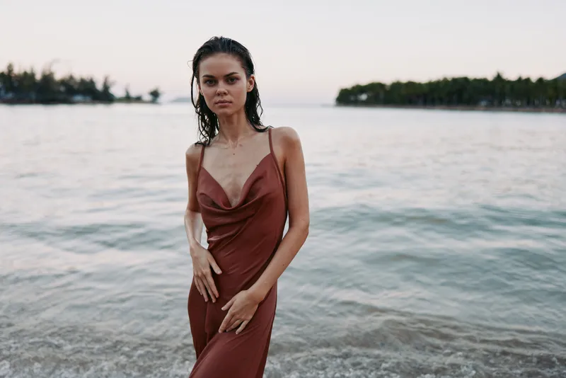 Elegant woman stands at the shoreline wearing a rust dress, calm expression, soft lighting, gentle waves, and a serene coastal portrait capturing poise and grace.