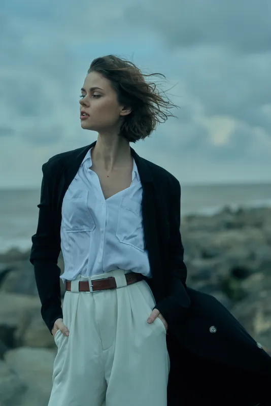 Woman in a smart blazer over a white shirt and wide pants stands outdoors by the windy coast, a flowing coat draped over her arm creating a bold fashion statement against a moody sea backdrop