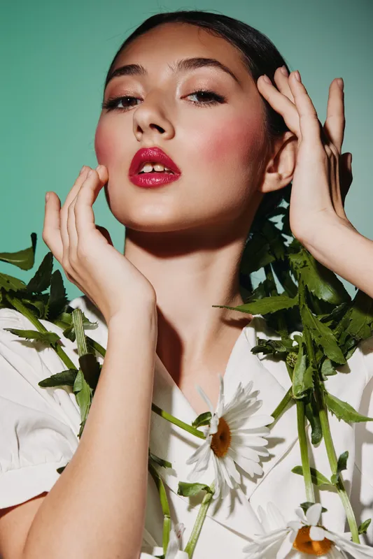 Close-up of young woman with vibrant glossy cranberry lips and natural soft makeup posing with green leaves and white daisies against green background. Concept of beauty fashion creative.