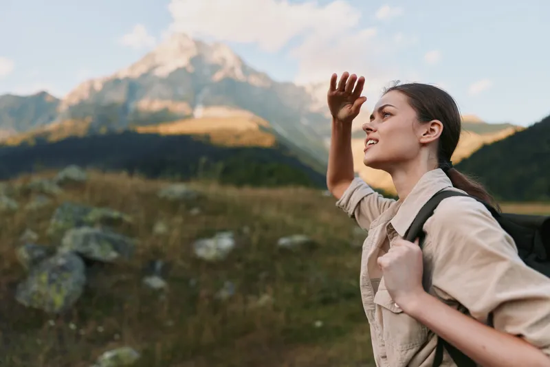 Young woman in a mountain meadow shields her eyes from the sun with a smile, backpack on, as distant peaks glow in golden light against a blue sky with wispy clouds nearby.