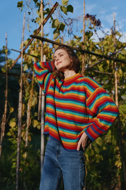 Woman in her mid 20s wearing a colorful striped sweater and jeans, smiling and leaning against a garden trellis among vines, relaxed lifestyle portrait in nature.
