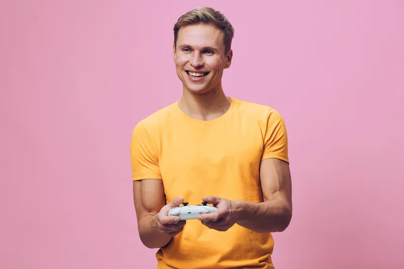 Smiling young man wearing a bright yellow t-shirt holding a game controller, looking happy and excited, standing against a plain pink background, studio portrait, lifestyle concept, joyful emotion, ca