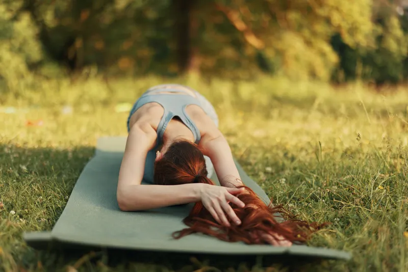 Young woman practicing yoga outdoors, wearing grey athletic wear, in a peaceful pose on a green mat surrounded by greenery, evoking tranquility and connection with nature
