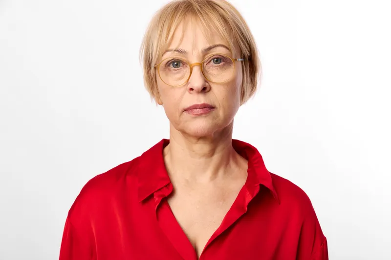 Woman senior portrait with glasses, red studio white background headshot, short blond hair and red shirt, calm expression, direct gaze, centered framing, soft studio lighting