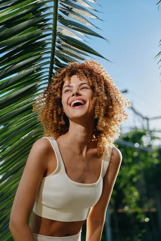 smiling woman with curly hair wearing white crop top outdoors under sunny sky by tropical palm leaves, joyful and relaxed, vibrant summer atmosphere