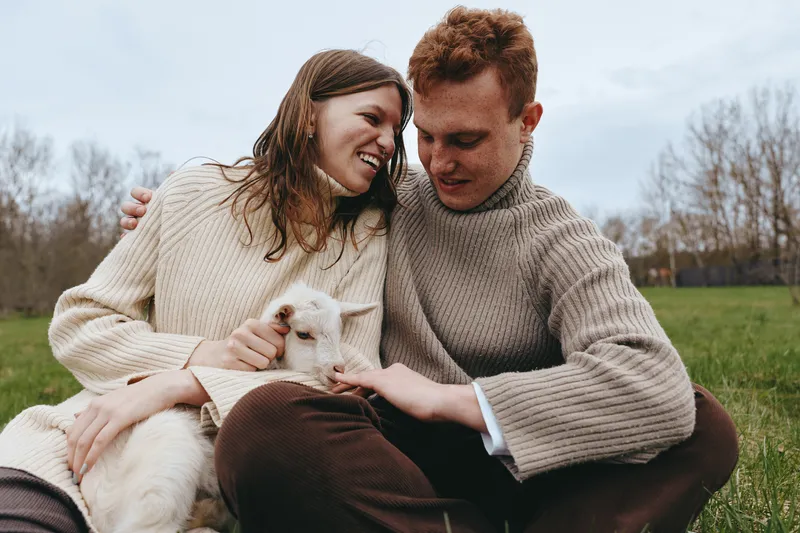 A smiling couple shares a gentle moment with a curious goat kid in an open field, showcasing warm affection, rural lifestyle, and simple happiness in a candid outdoor scene with nature.