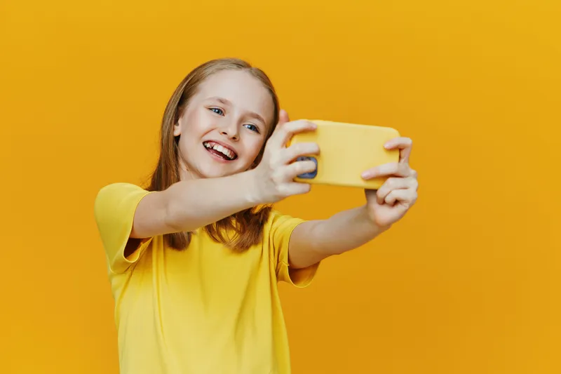 Cheerful girl taking a selfie with a yellow smartphone against a vibrant yellow background, showcasing joy, summer vibes, and bright colors