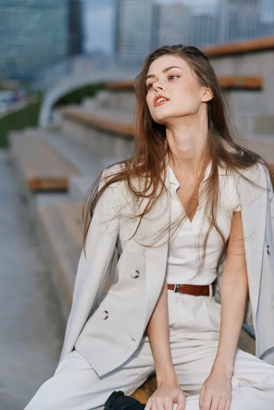 Confident young woman in beige suit jacket sitting outdoors on urban steps, stylish casual fashion pose with soft natural light, modern city background