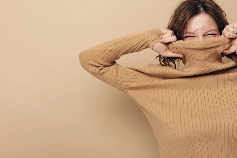Young woman in brown sweater pulls collar over face with eyes closed, expressing playful mood on beige background. Concept of casual fashion, emotions, and cozy style.