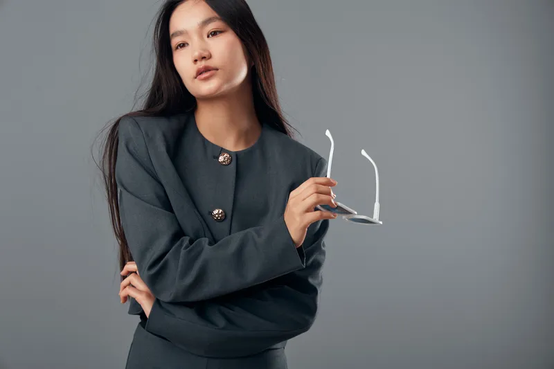 Young woman in gray blazer holding eyeglasses posing thoughtfully against gray background. Female model with long dark hair, confident expression, and stylish business attire in studio.
