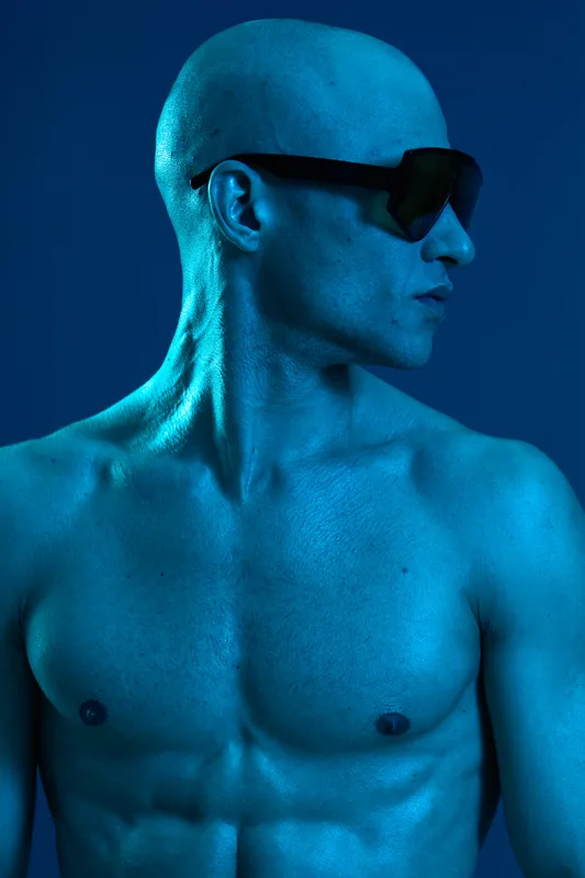 Muscular man with a shaved head wearing sunglasses, showcasing a striking blue hue, exuding confidence and a cool vibe, against a solid background.