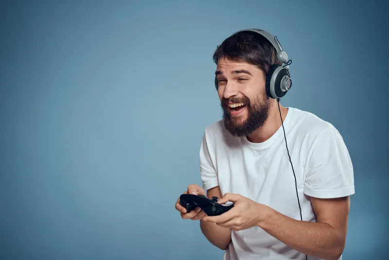 Мужчина в наушниках, контроллер в руках, играет в игровую консоль, образ жизни, эмоции, голубой фон, белая футболка