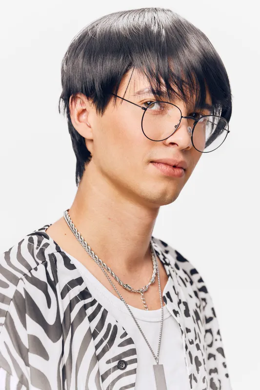 young man with glasses wearing patterned shirt and layered chains looks confidently at camera with neutral white background, trendy casual style in natural light