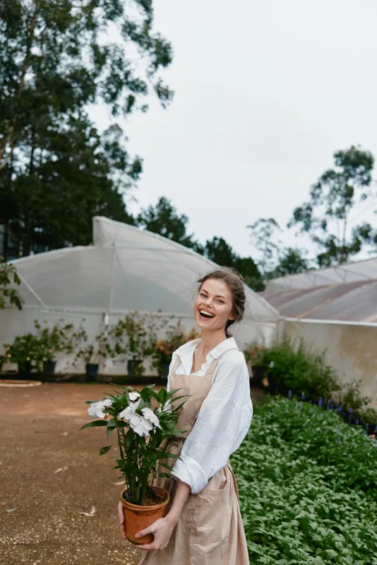 Joyful young woman holding a potted plant in a lush greenhouse, wearing a white shirt and beige apron, surrounded by greenery and natural lighting