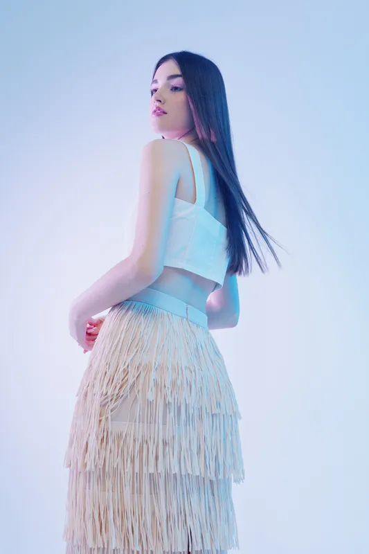 Young woman with long dark hair wearing a white top and fringed skirt poses against a light background. Concept of woman beauty fashion and style.