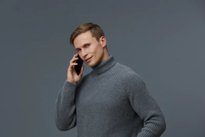Smiling man talking on mobile phone, wearing cozy gray turtleneck sweater, modern casual style, confident and relaxed mood, neutral background, studio portrait, lifestyle concept.
