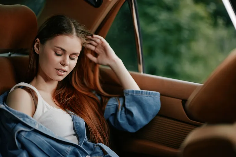 Young woman with long, auburn hair in a denim jacket, looking thoughtful while sitting in a car, surrounded by lush green background, evoking a sense of tranquility.