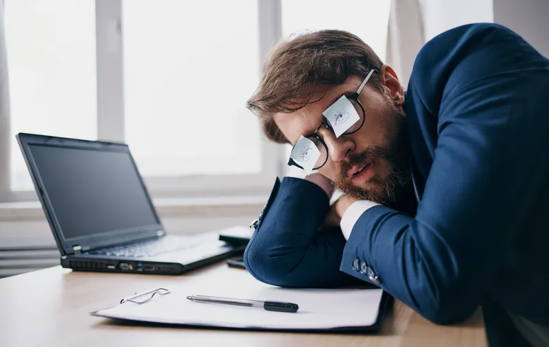 Бизнесмен в очках спит на работе, рядом с ним разложены листы бумаги