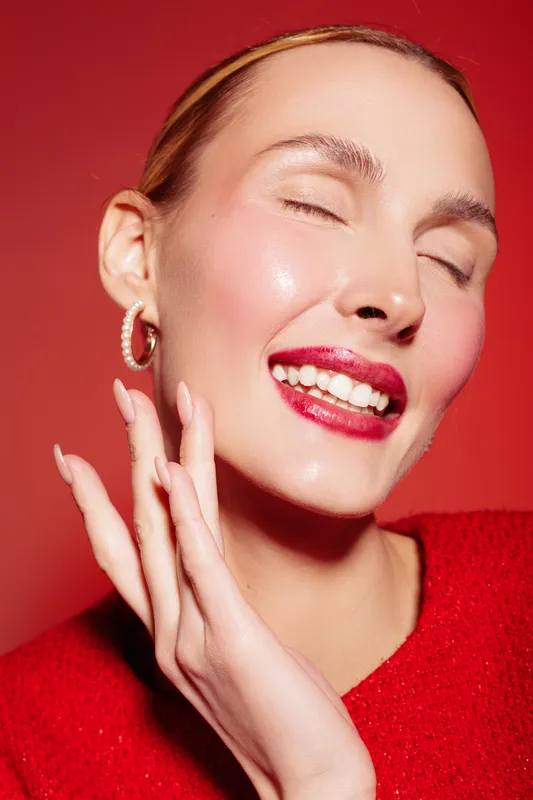 Close-up of smiling woman with red lipstick and glowing skin wearing red sweater and hoop earrings. She poses with closed eyes and hand near face on red background. Beauty and makeup concept.