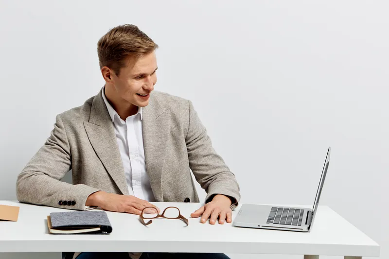Professional young man in formal blazer sitting at desk with laptop and glasses on white background, working and smiling, business casual style, office work, technology. People lifestyle concept