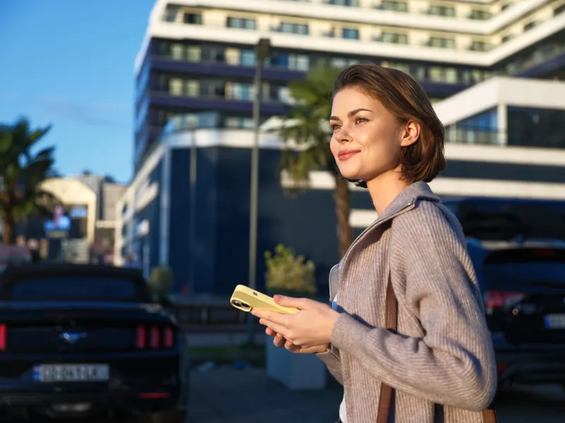 A confident female professional stands in a modern outdoor setting, holding a smartphone and smiling, projecting a bright, engaged mood and contemporary business style.