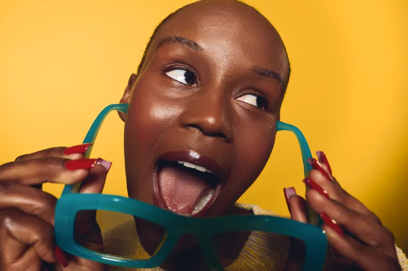 Close-up of a happy dark skinned African woman with short hair holding oversized blue glasses near her mouth on yellow background. Concept of joy, fun, and playful fashion portrait.
