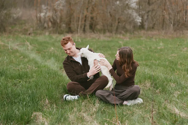 Cheerful couple outdoors, sharing a moment with a friendly goat kid, gentle laughter and warm light, relaxed pose, pastoral grass setting, soft breeze, natural colors, wholesome lifestyle