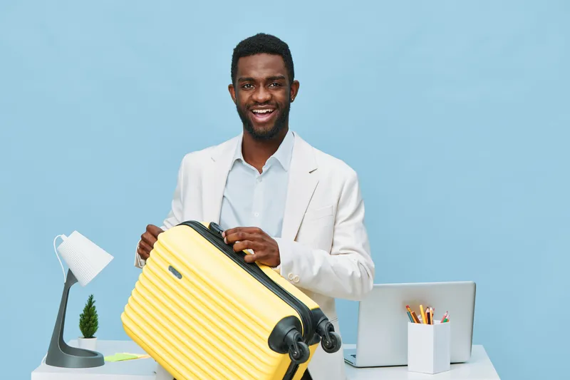 Smiling man with dark skin dressed in white suit jacket holding yellow suitcase in modern office with laptop, desk lamp, stationery and blue background. Travel, business trip, professional concept.