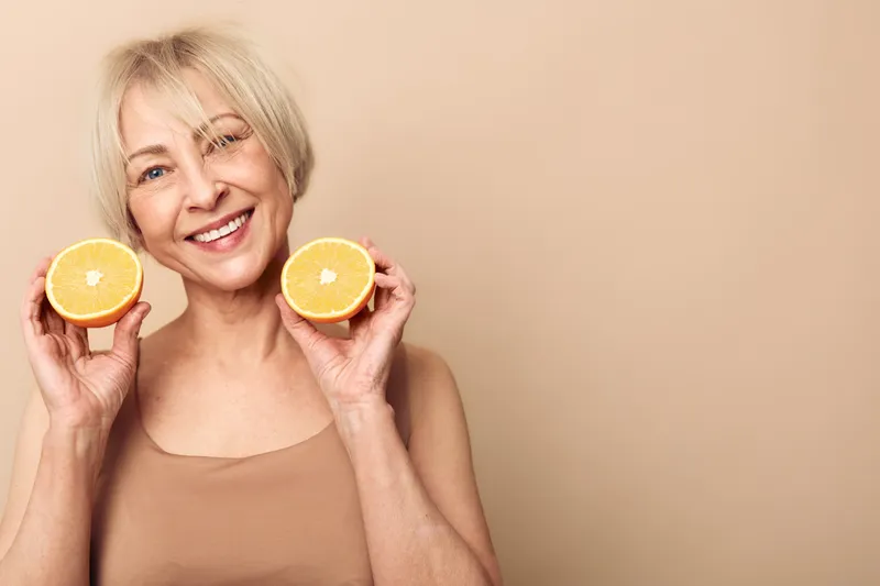 Smiling mature woman with short blonde hair holds two halves of fresh orange near face on beige background. Concept of healthy lifestyle, vitamin C, natural beauty, and happiness.