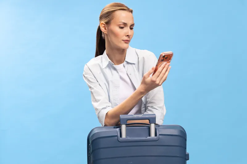 Young woman with suitcase looks at smartphone screen with serious expression on blue background. Concept of travel, waiting, communication, and planning before trip.