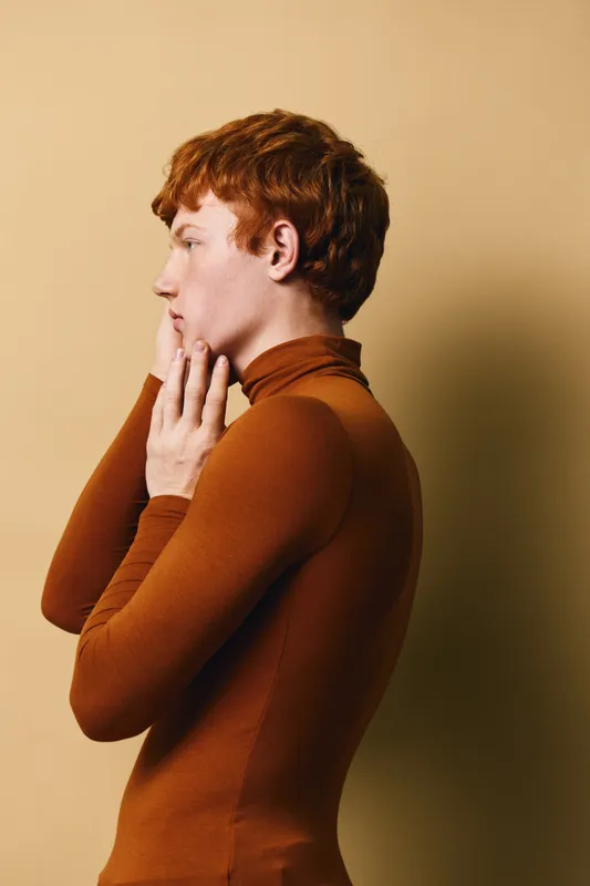 Profile of young man with red hair and pale skin wearing brown turtleneck sweater standing against beige background looking thoughtful and contemplative.