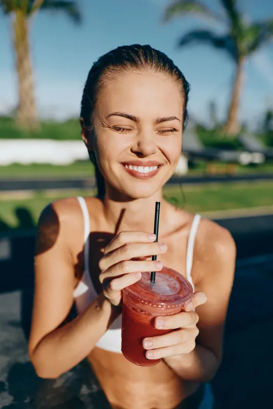 Bright woman in a bikini by the pool drinks a red smoothie with a straw, enjoying a sunny day, relaxed lifestyle, cheerful mood, outdoor refreshment, tropical poolside setting.