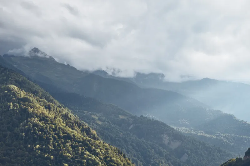 Majestic mountain landscape with lush slopes and misty clouds drifting over a tranquil valley, offering a wide panoramic view of rugged ridges, dense forest, and expansive terrain under a moody sky.