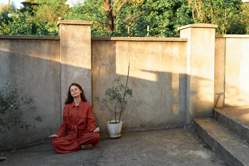 A woman in a red dress sits cross-legged on a concrete ground beside a weathered wall in bright outdoor light, creating a calm, contemplative lifestyle moment