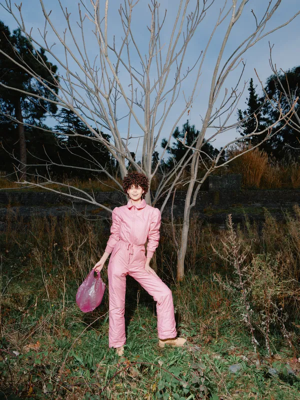 Fashion woman pink jumpsuit outdoor portrait with bag beside a bare tree, natural scene and bold color, showcasing confident pose and lifestyle style for editorial fashion photography
