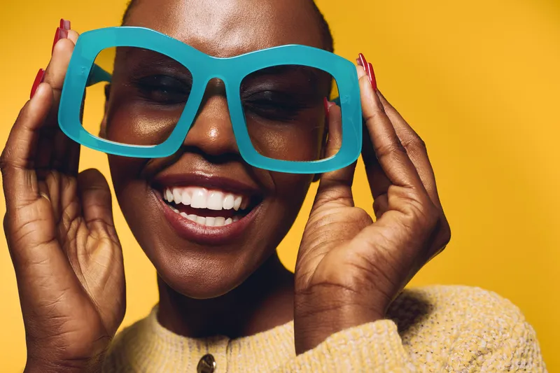 Smiling dark skinned African woman wearing large blue glasses and yellow sweater posing on yellow background. Concept of happiness, fashion, and joyful lifestyle with vibrant colors.