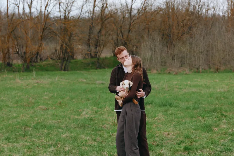 Young couple embraces tenderly in a green field while holding a small baby goat, enjoying a peaceful moment in nature during a casual autumn day.