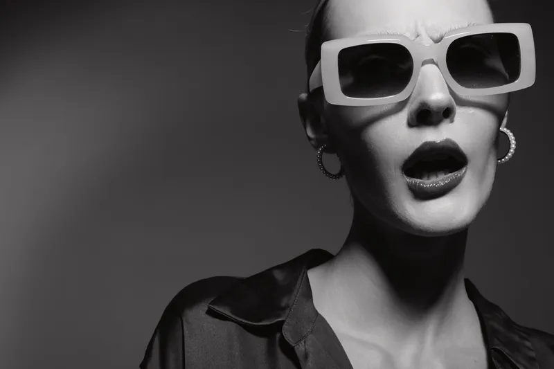 Portrait of a pale white woman with sunglasses and hoop earrings in black blouse shows surprised expression in studio lighting.