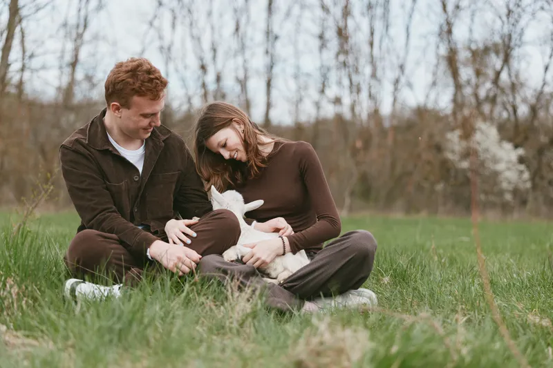Couple sits on grass outdoors, holding a goat kid with affection, sharing smiles in a serene countryside park, a wholesome lifestyle moment of nature and family warmth.