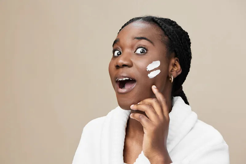 Young Black woman applying skincare cream, surprised expression, in a white bathrobe against a neutral beige background, perfect for beauty and skincare themes