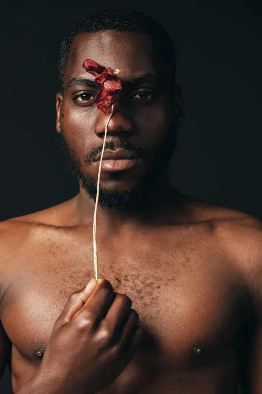 Black man with bare chest holding dried red flower in front of face, creative conceptual portrait with visual style, artistic expression and dark background for emotional impact.